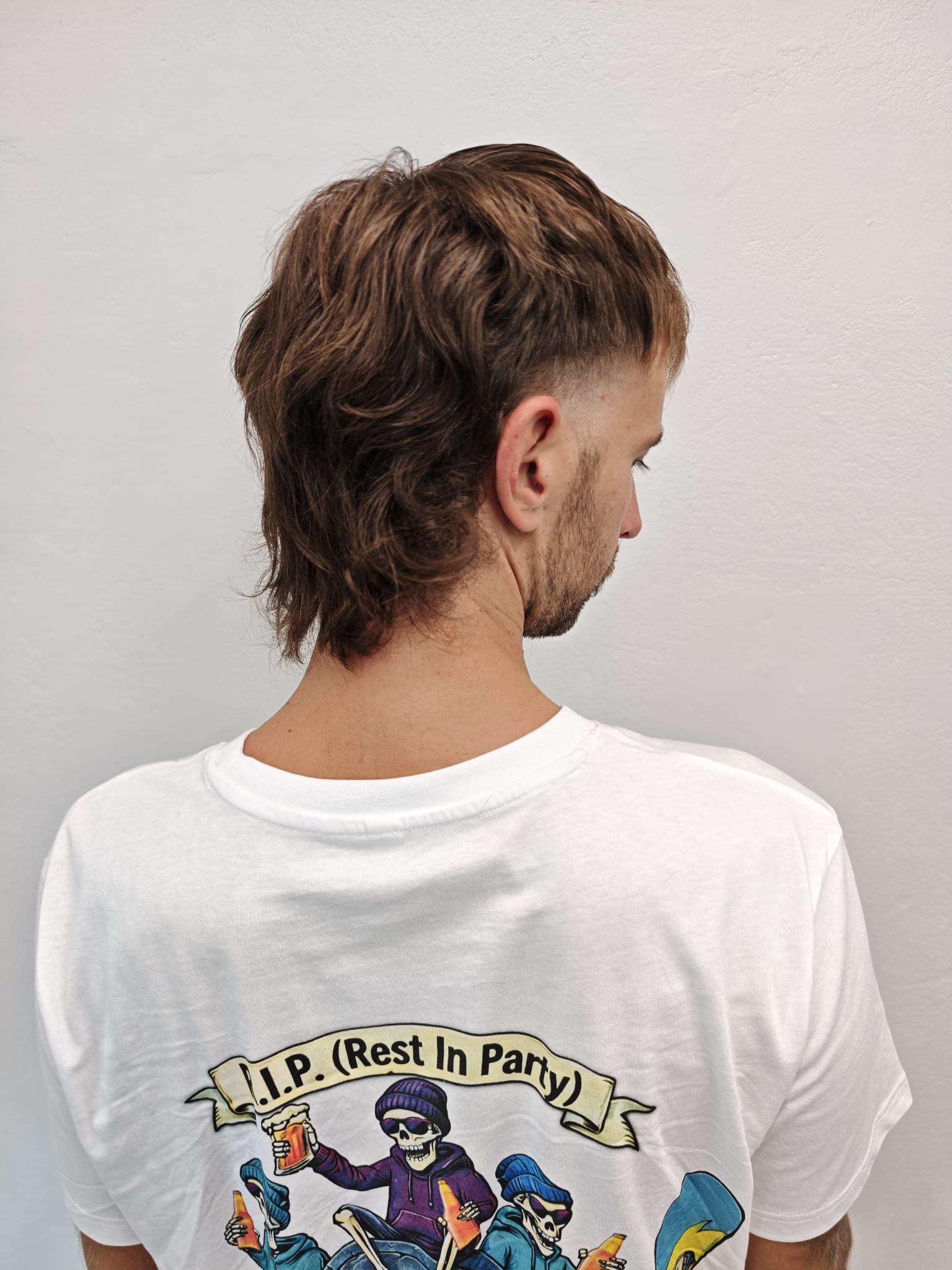 Profile of a young man with curly brown hair and light facial stubble wearing a black shirt against a plain light gray background.