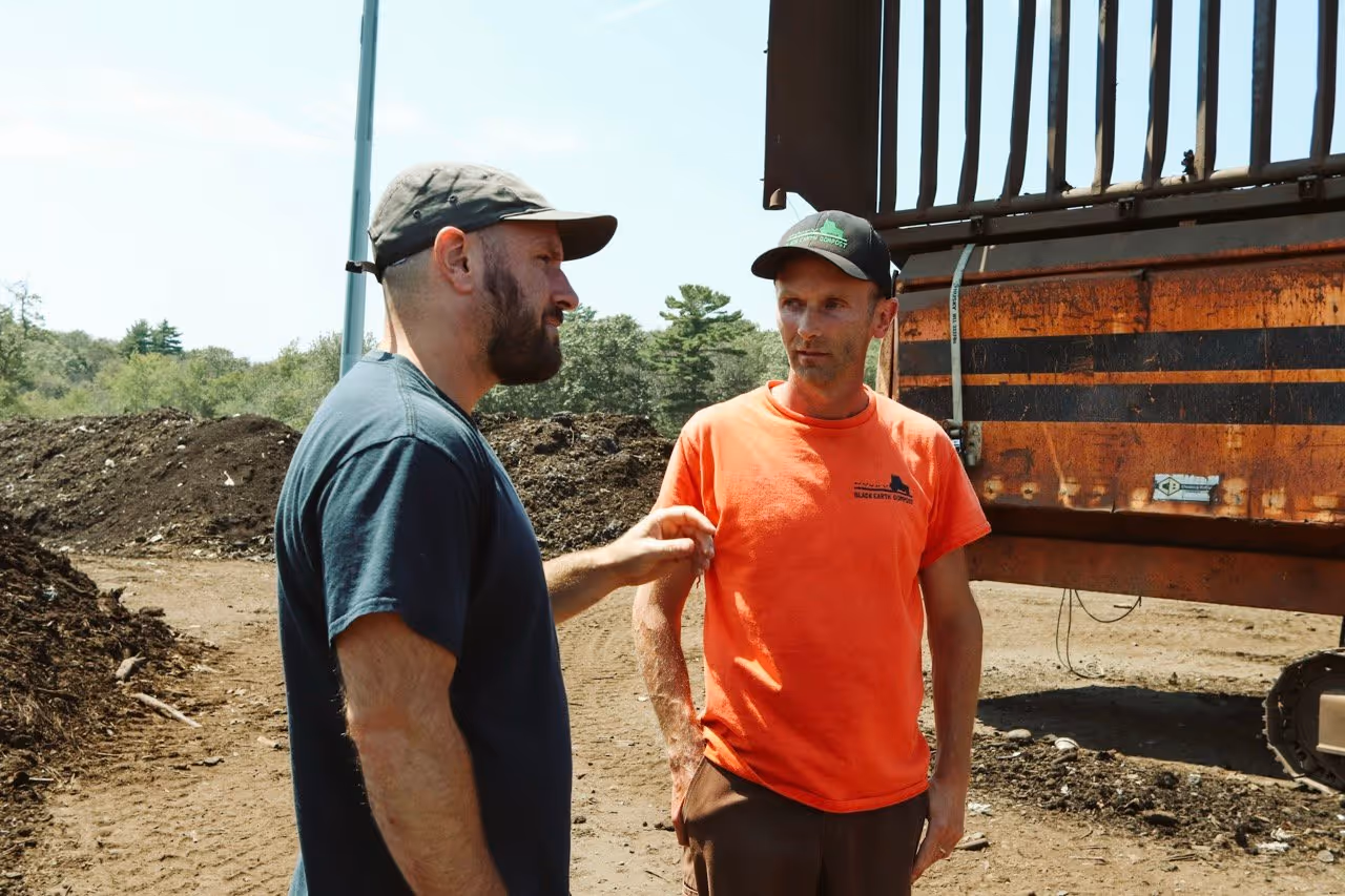 Two men having a conversation outdoors near piles of soil and heavy machinery at a compost site.