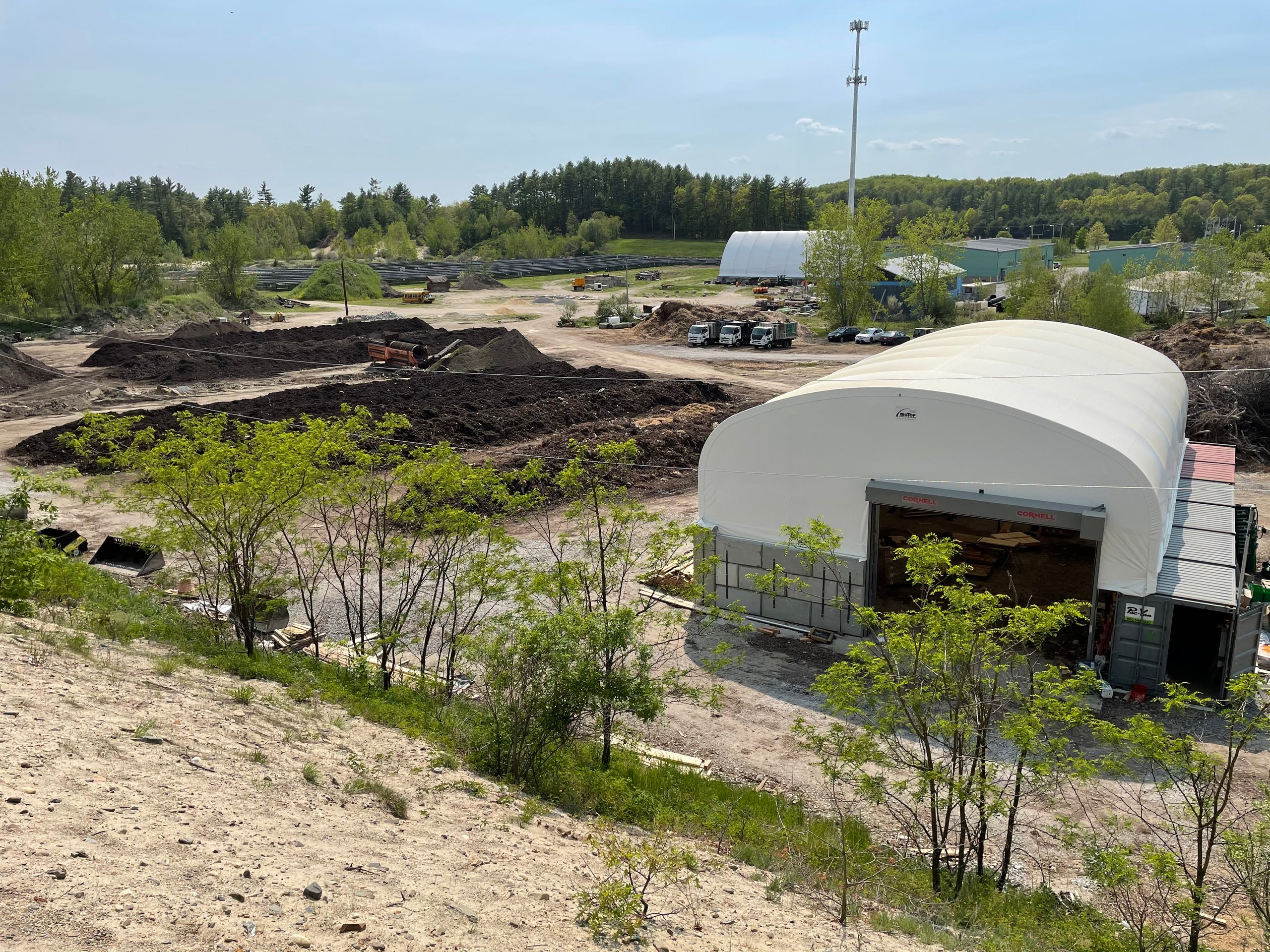 Compost facility with large piles of soil and organic material, white industrial tents, trucks, and surrounding green trees under a clear sky.