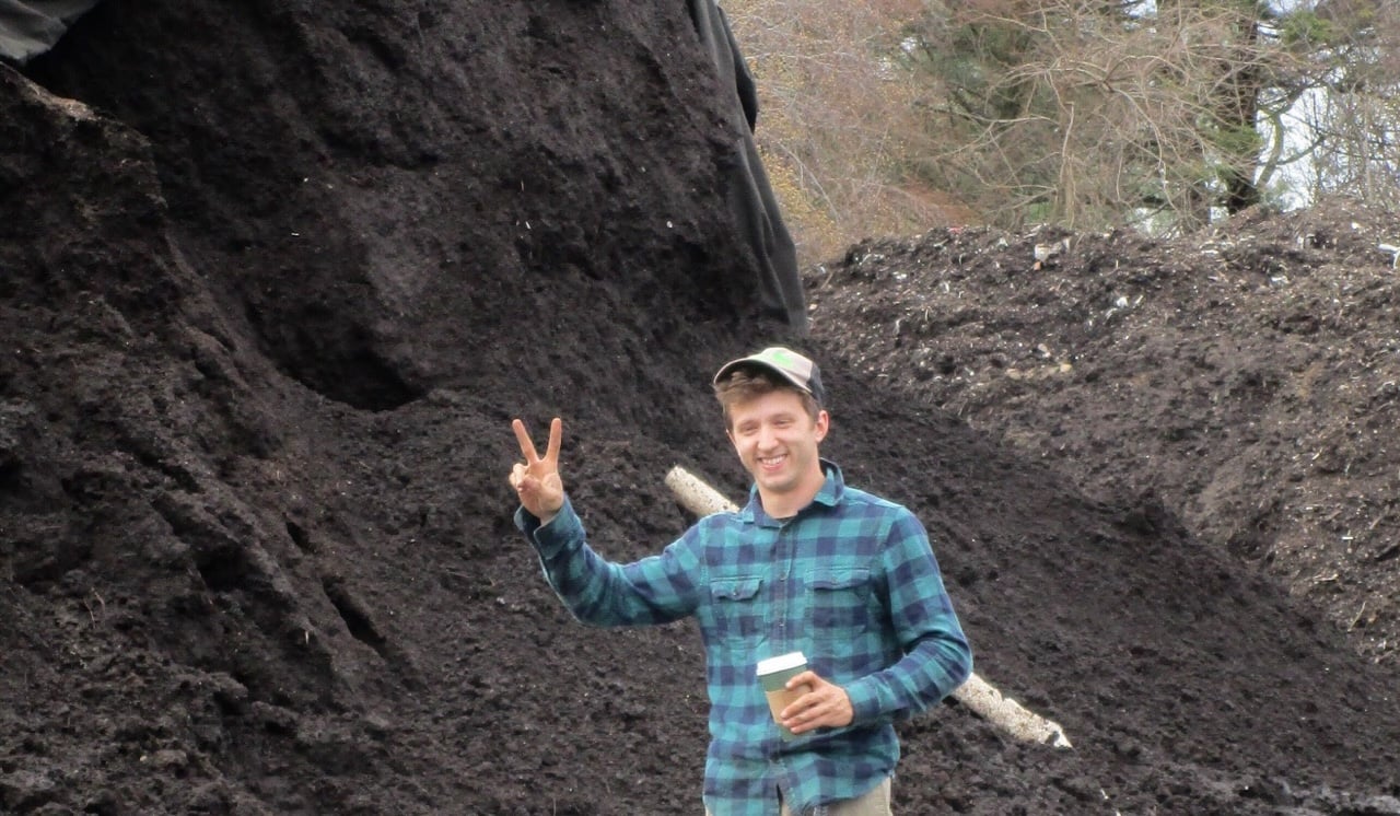 Man in blue plaid shirt and cap holding a coffee cup and making a peace sign gesture in front of large piles of dark soil or compost outdoors.