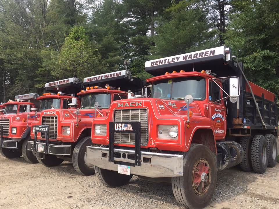 Row of red Mack dump trucks with Bentley Warren signage parked on gravel in front of green trees.