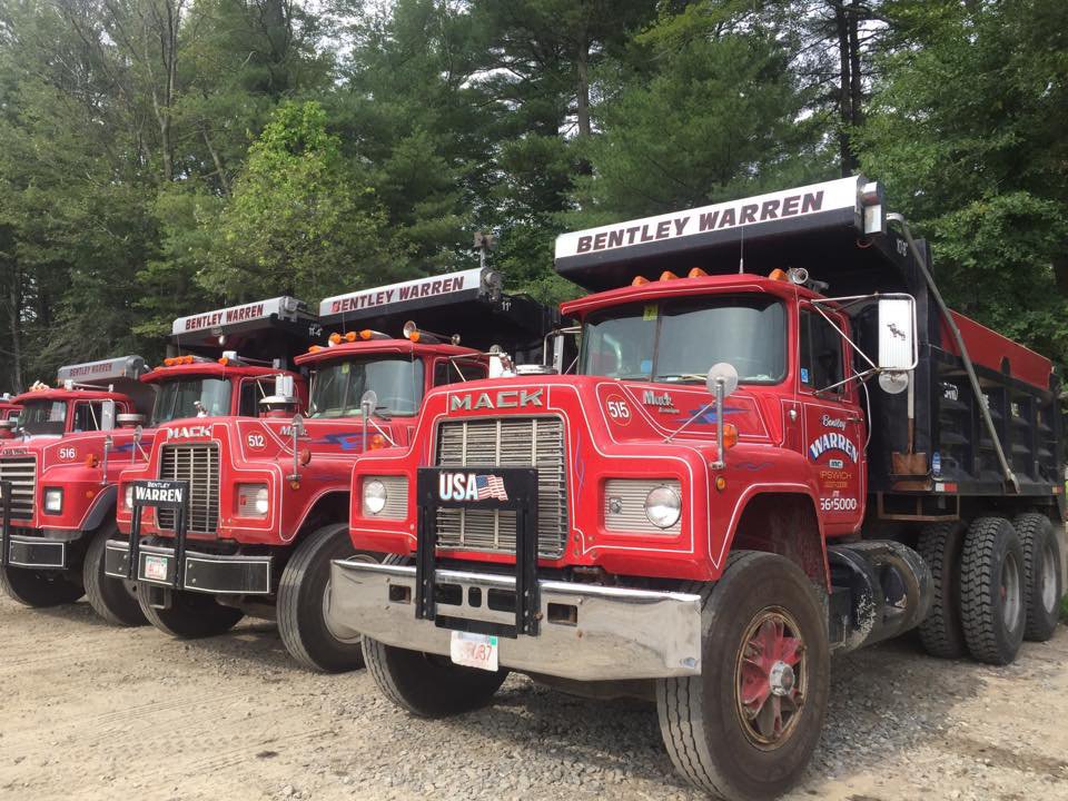 Row of red Mack dump trucks with Bentley Warren signage parked on gravel in front of green trees.