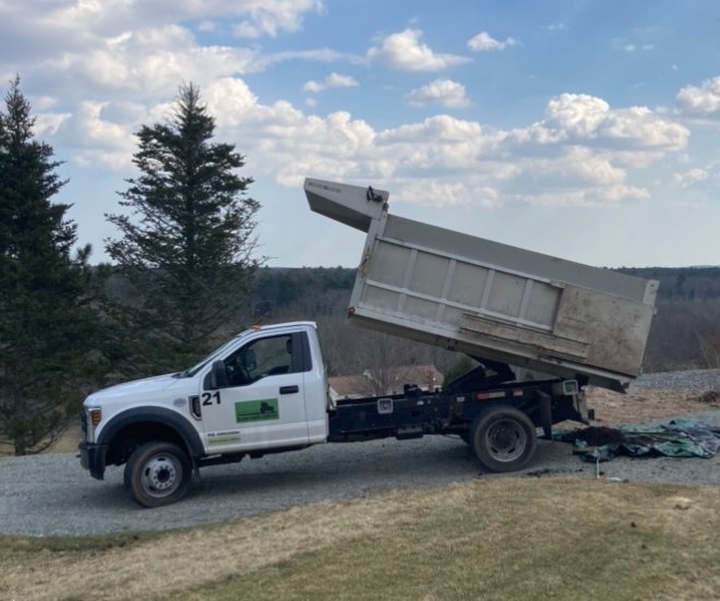 White dump truck with raised bed parked on gravel driveway near grassy yard with trees and cloudy sky in background.