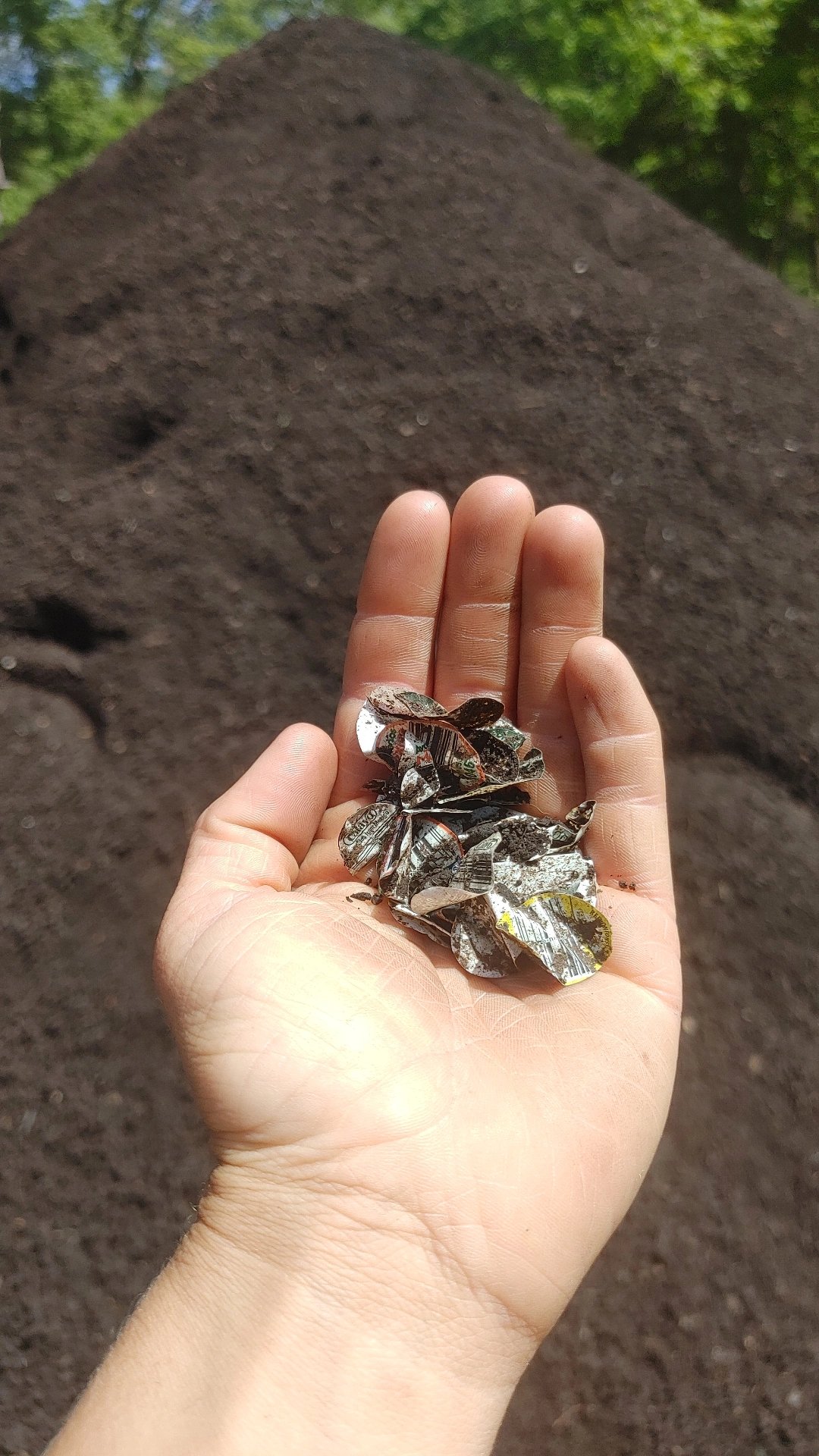 Hand holding small pieces of soil-covered metallic or foil debris with a pile of dark soil in the background.