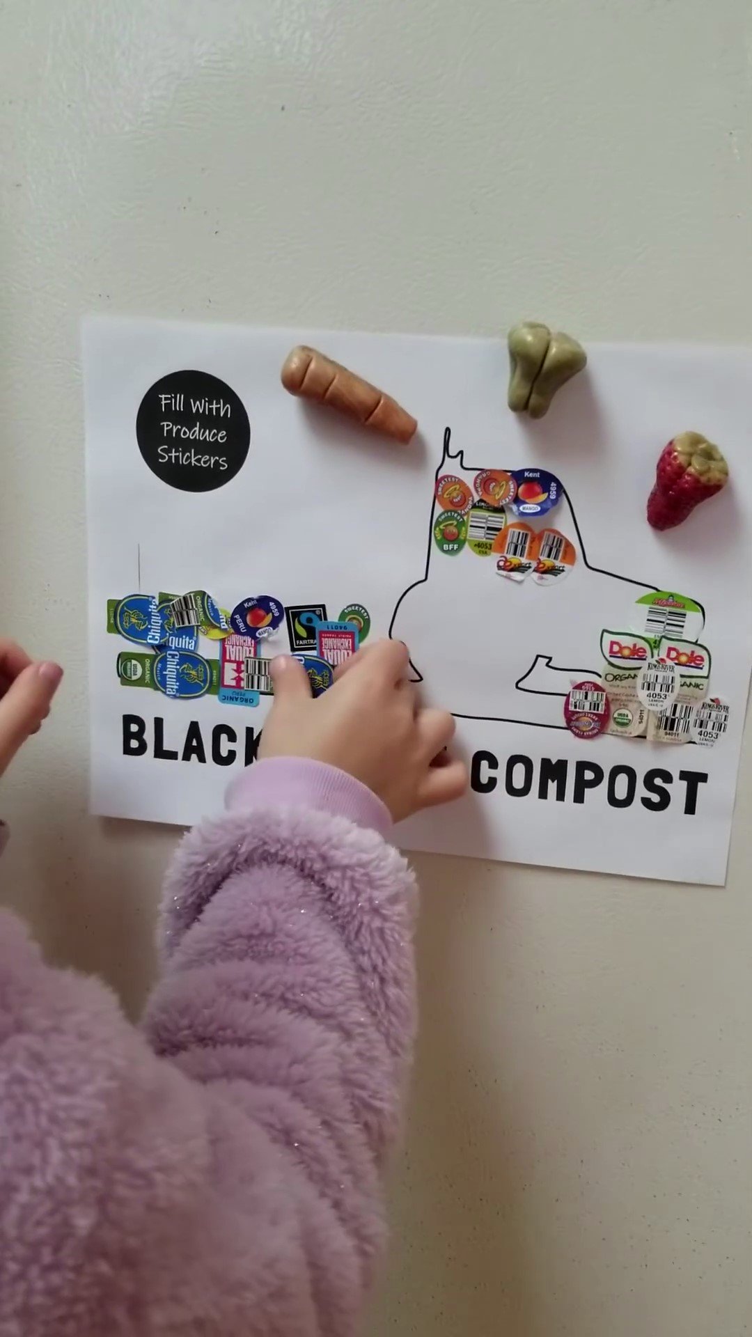 Child placing fruit stickers on a black compost poster shaped like a cat, with additional small toy vegetables attached at the top.