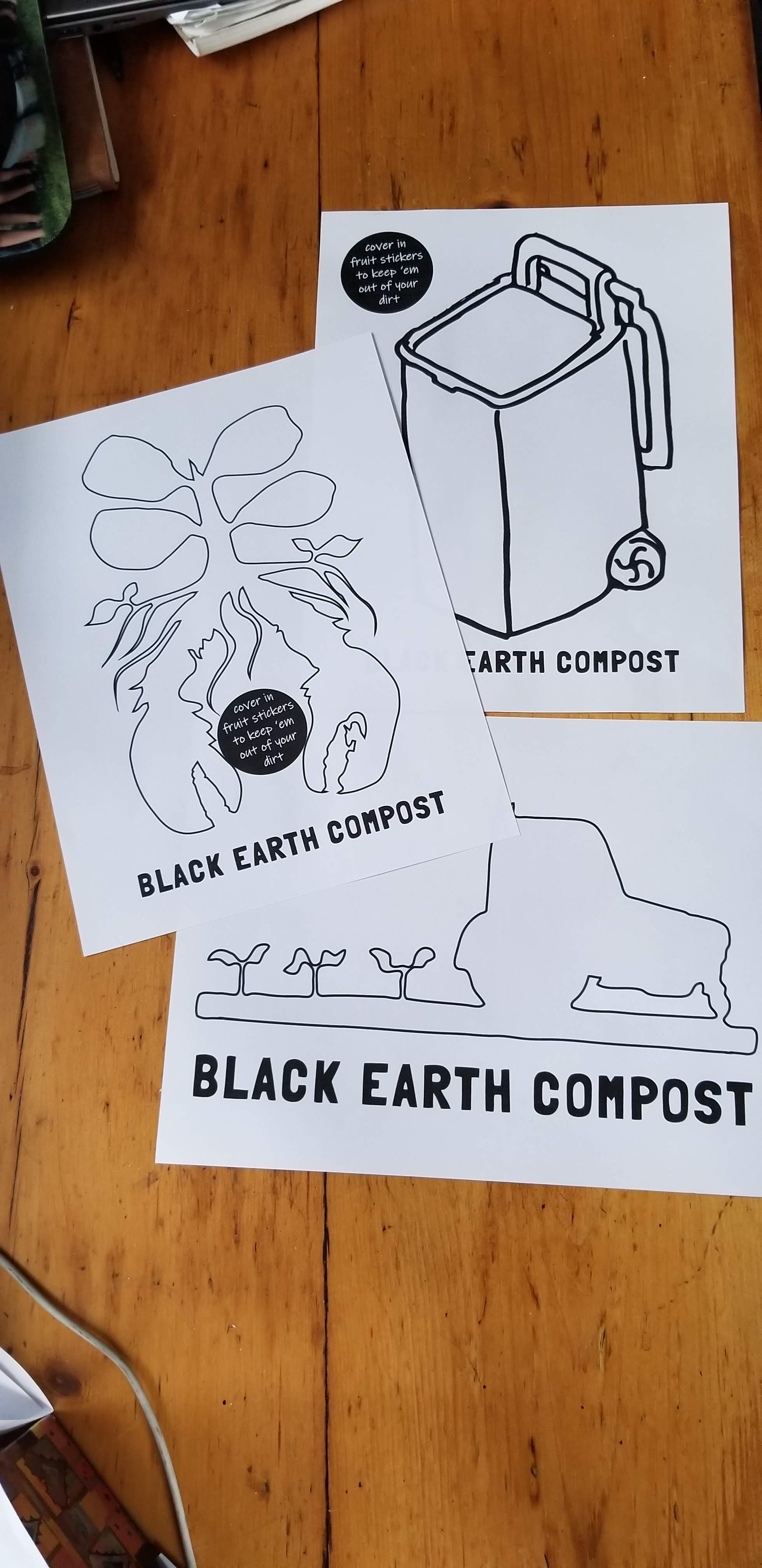 Three Black Earth Compost flyers on a wooden table featuring simple black line drawings of a compost bin, a plant with roots and a lobster claw, and a farming tractor with seedlings, each with the text 'BLACK EARTH COMPOST' and a note about covering in fruit stickers to keep out dirt.