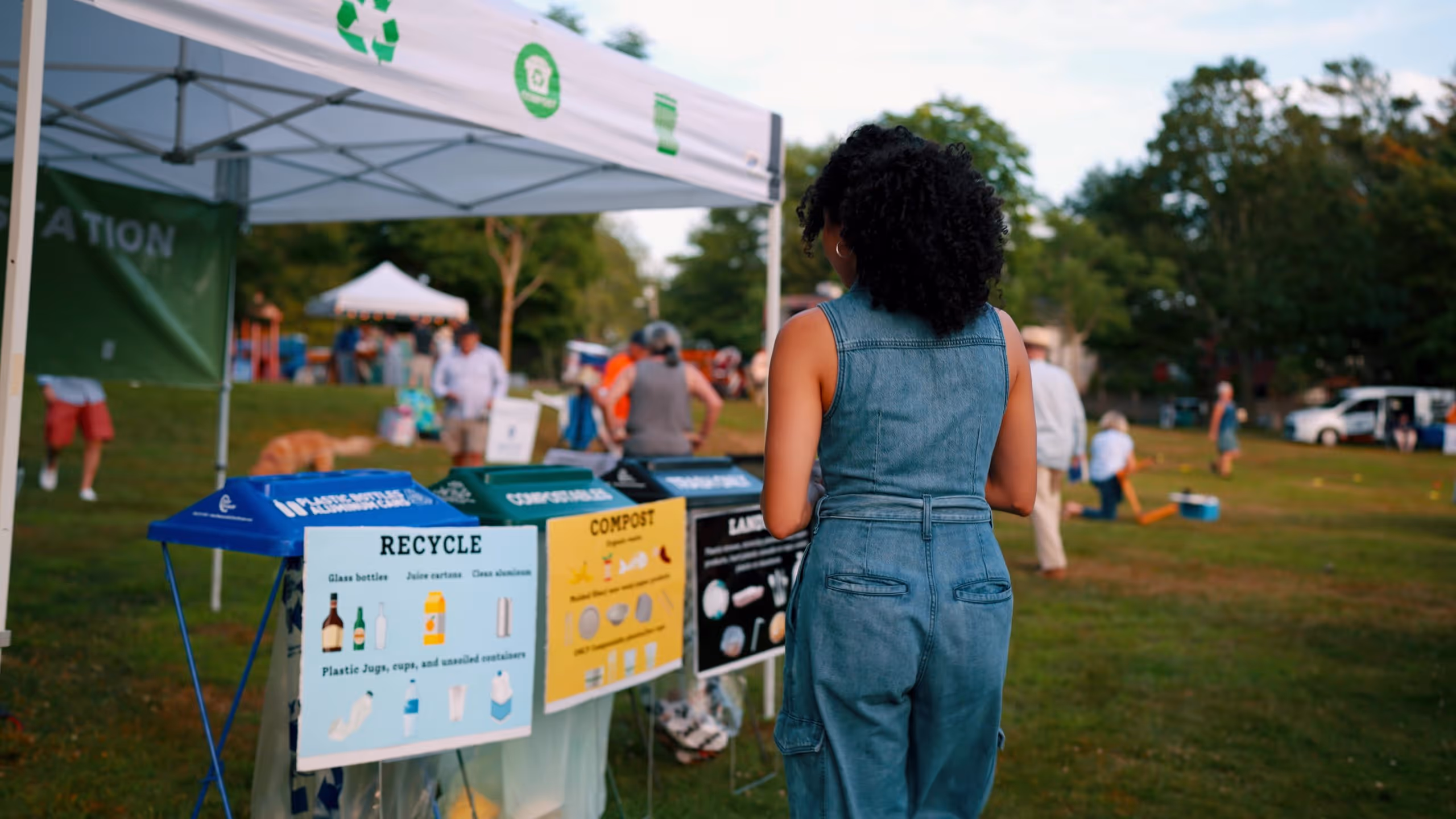 Woman in denim jumpsuit standing near recycling, compost, and landfill bins at an outdoor event with people and trees in the background.