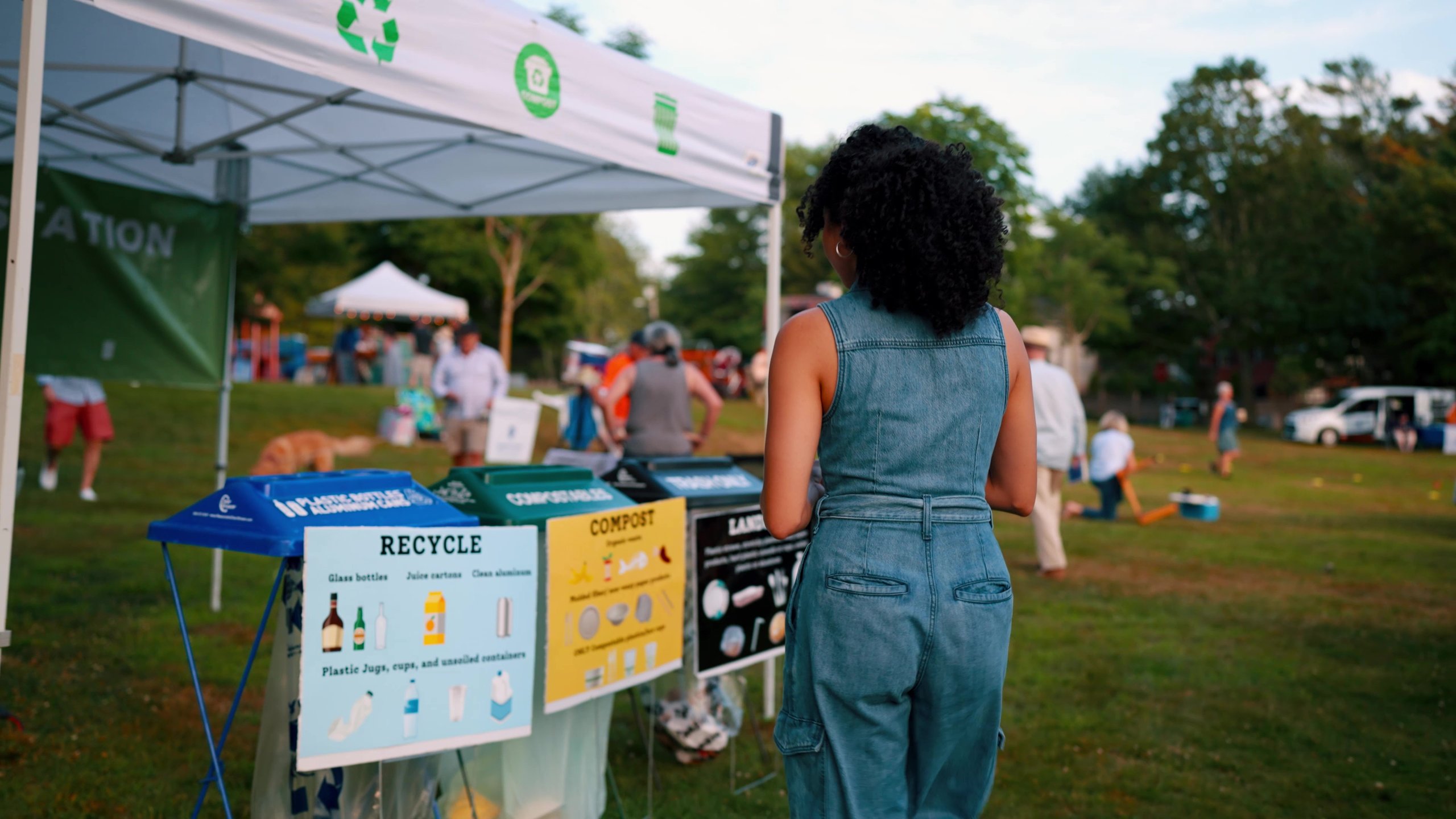 Woman in denim jumpsuit standing near recycling, compost, and landfill bins at an outdoor event with people and trees in the background.