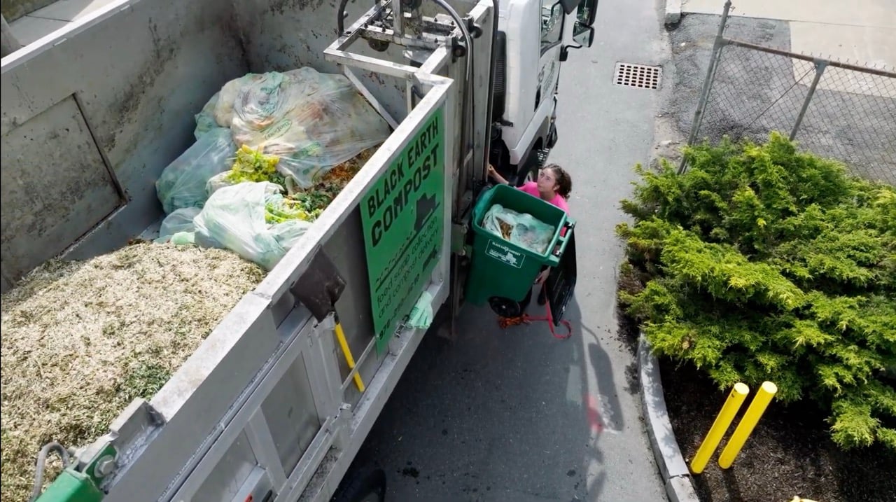 Person in a pink shirt emptying a green compost bin into a Black Earth Compost truck filled with organic waste.