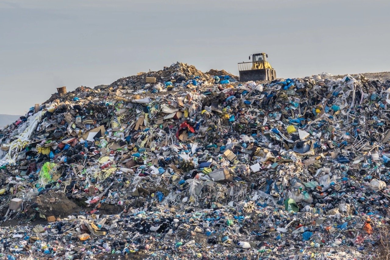 Large landfill pile with scattered garbage and a bulldozer on top under a cloudy sky.