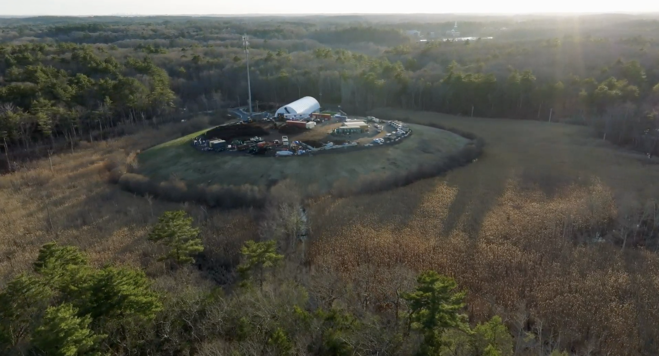 Aerial view of a circular clearing with a construction site and vehicles surrounded by dense forest and fields under sunlight.