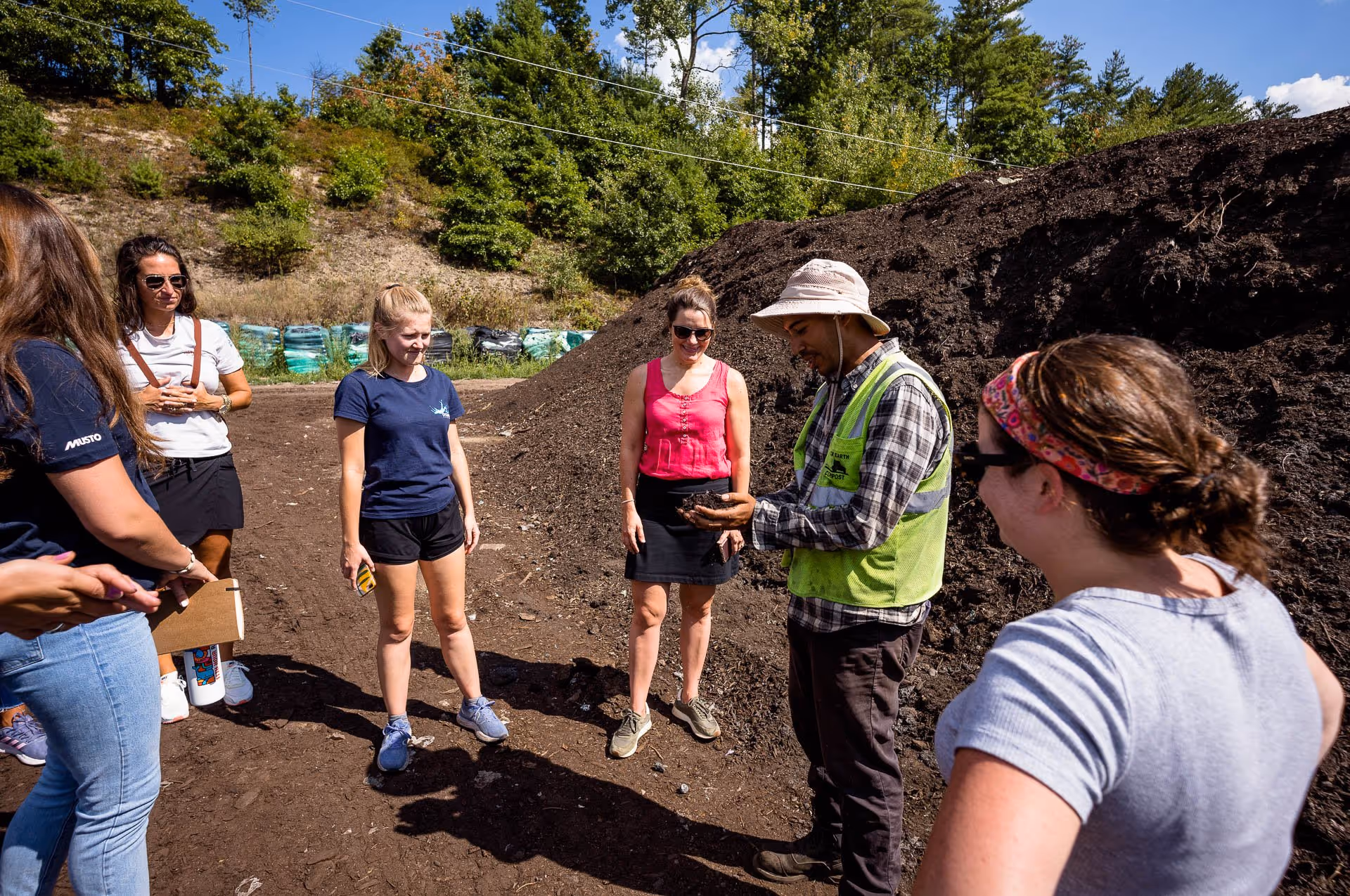 Group of six people outdoors standing on dirt near a large compost pile; one man in a vest and hat shows soil to the others.