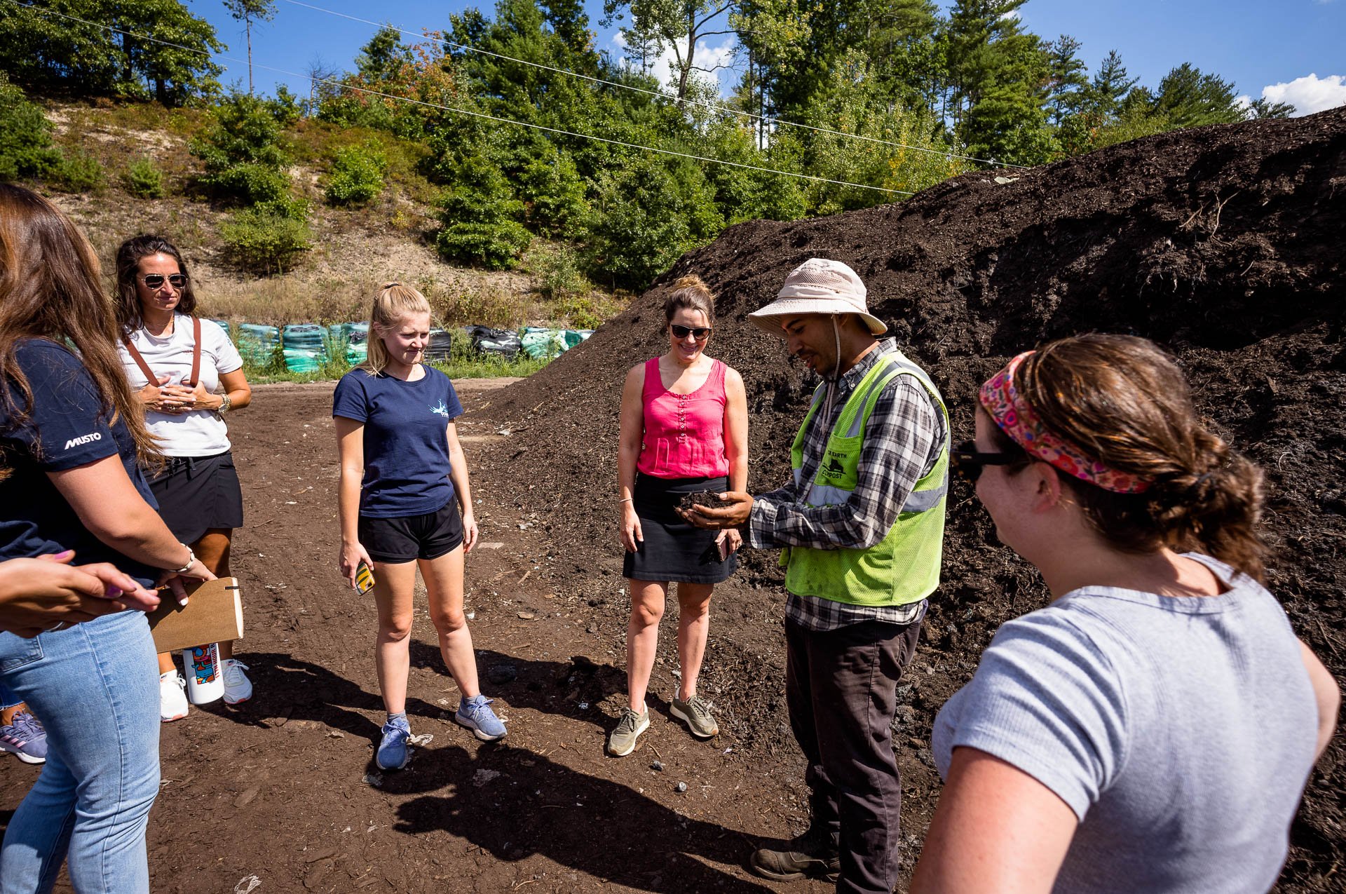 Group of six people outdoors standing on dirt near a large compost pile; one man in a vest and hat shows soil to the others.