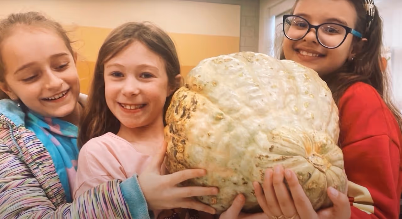 Three smiling girls holding a large, light-colored pumpkin indoors.
