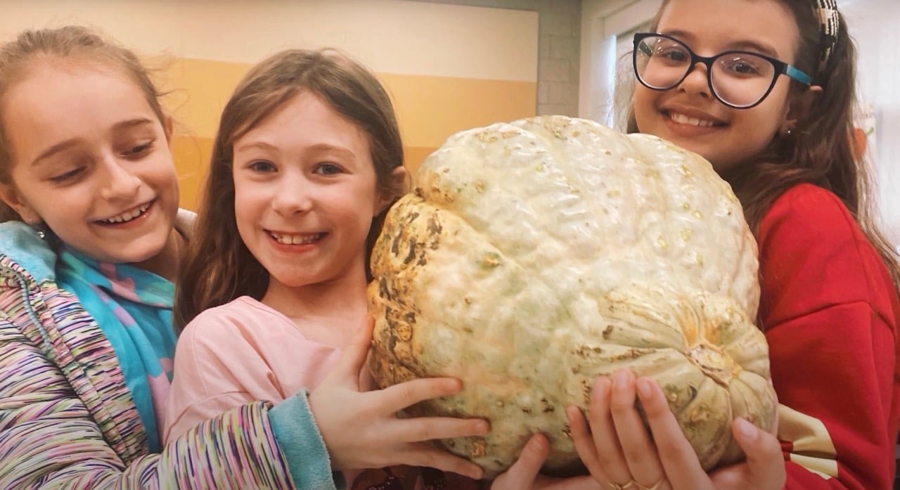 Three smiling girls holding a large, light-colored pumpkin indoors.