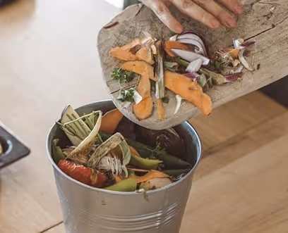 Hand emptying vegetable scraps including onion peels and sweet potato skins into a metal compost bucket.