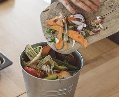 Hand emptying vegetable scraps including onion peels and sweet potato skins into a metal compost bucket.