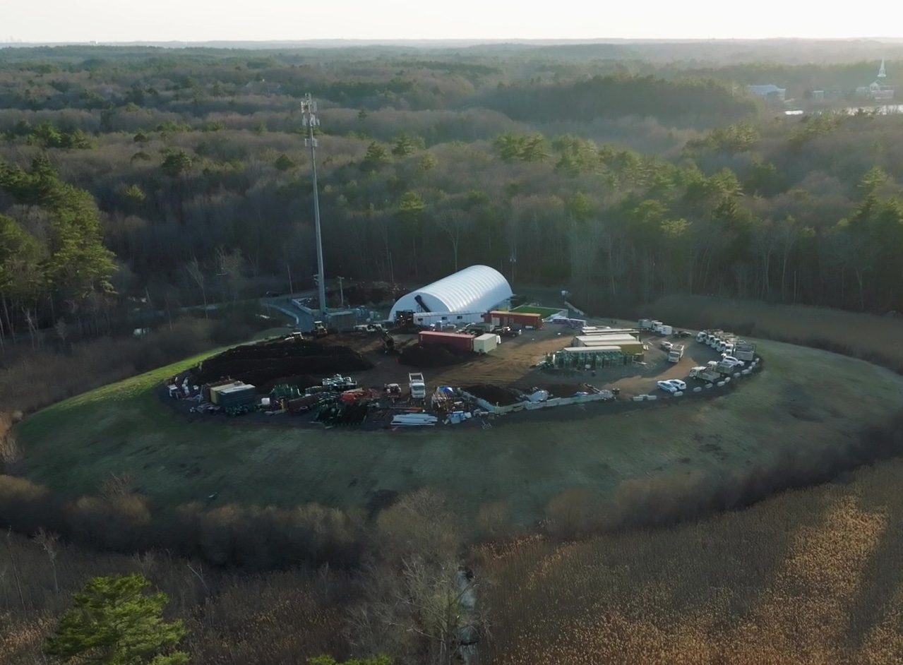 Aerial view of a fenced construction or industrial site with a large white arched building, heavy machinery, and trucks, surrounded by dense forest.