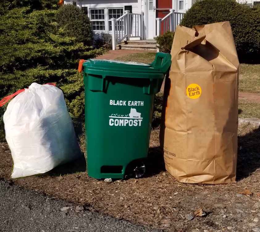 Green compost bin labeled 'Black Earth Compost' next to a white plastic bag and a large brown paper yard waste bag with 'Black Earth' sticker.