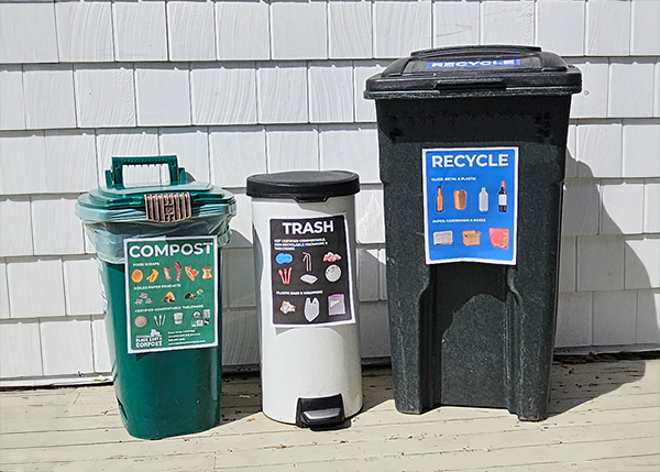 Three labeled bins for compost, trash, and recycling lined up outdoors against a white wooden wall.