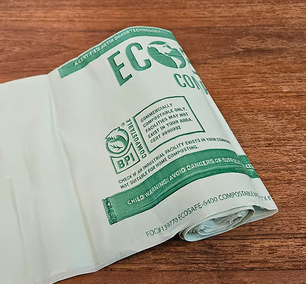 Rolled white eco-friendly biodegradable compostable bag with green text and certification logos on a wooden surface.