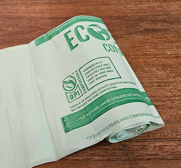 Rolled white eco-friendly biodegradable compostable bag with green text and certification logos on a wooden surface.