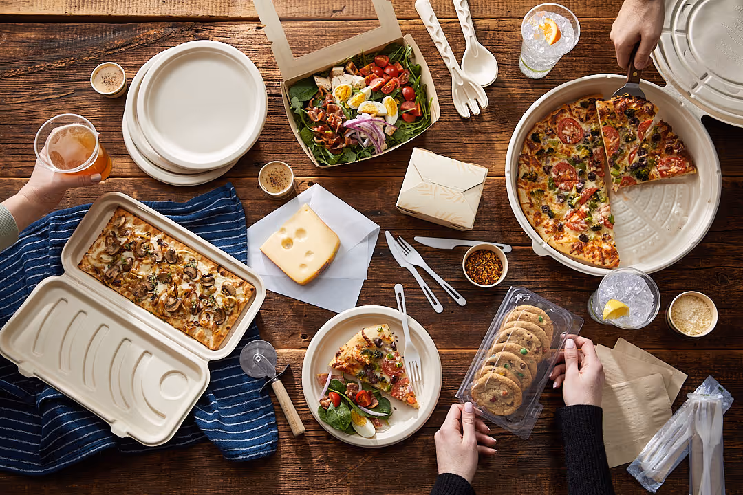 Overhead view of a wooden table with assorted food including two pizzas, a salad, a flatbread with mushrooms, a block of cheese, cookies in plastic packaging, drinks, and eco-friendly disposable plates and utensils.