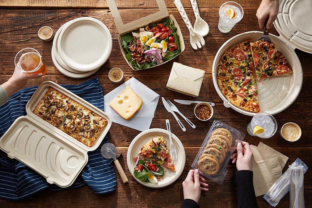Overhead view of a wooden table with assorted food including two pizzas, a salad, a flatbread with mushrooms, a block of cheese, cookies in plastic packaging, drinks, and eco-friendly disposable plates and utensils.