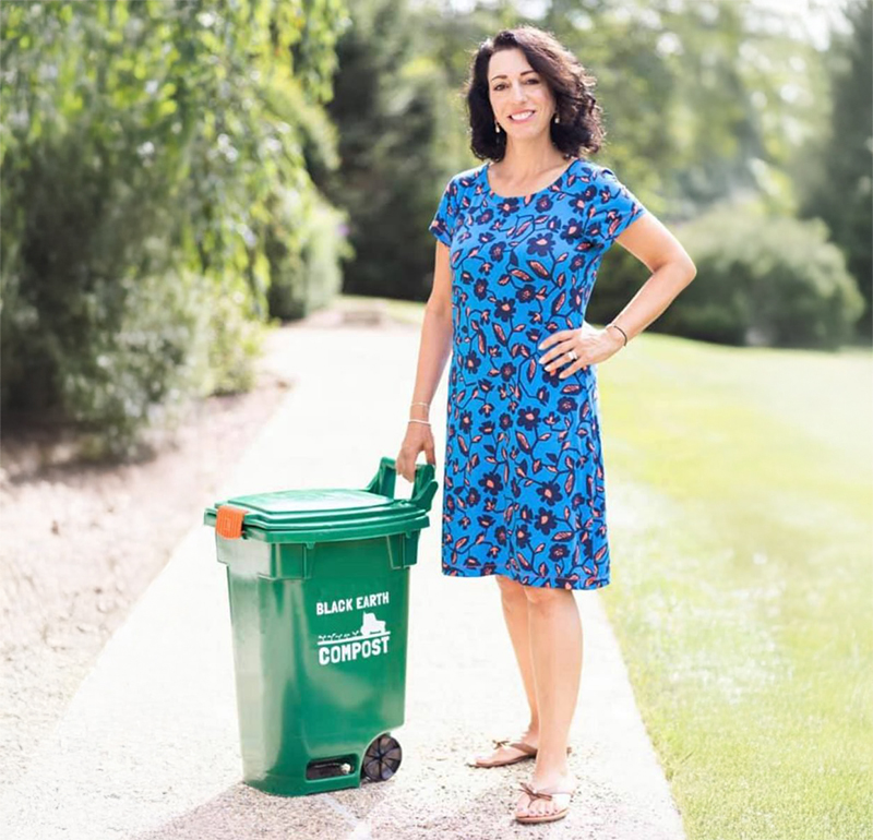 Woman in a blue floral dress standing outdoors on a path, holding a green compost bin labeled 'Black Earth Compost'.
