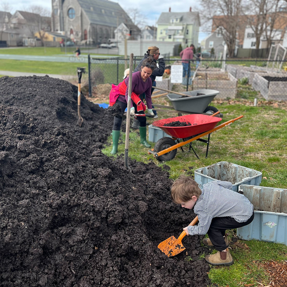 A young boy shoveling soil from a large pile into containers while adults with wheelbarrows work in a community garden near a church and houses.