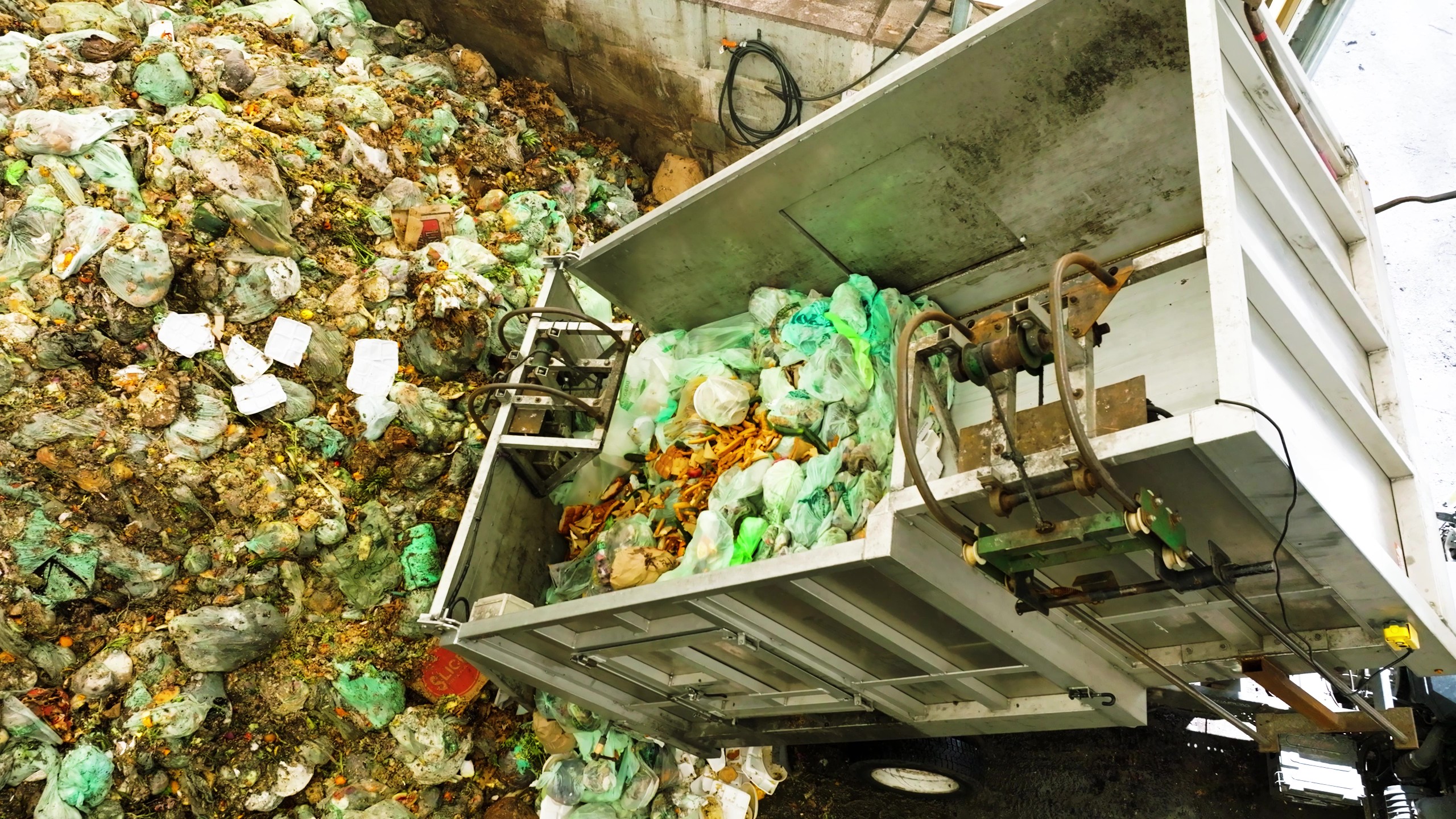 Garbage truck unloading bags of waste into a large indoor waste collection area filled with piled trash bags and food scraps.