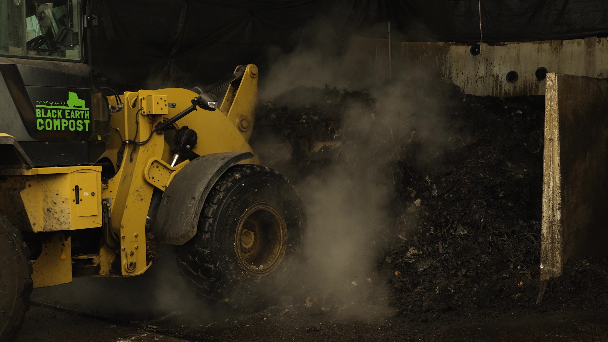 Yellow loader machine mixing steaming compost inside a building with Black Earth Compost logo on the vehicle.