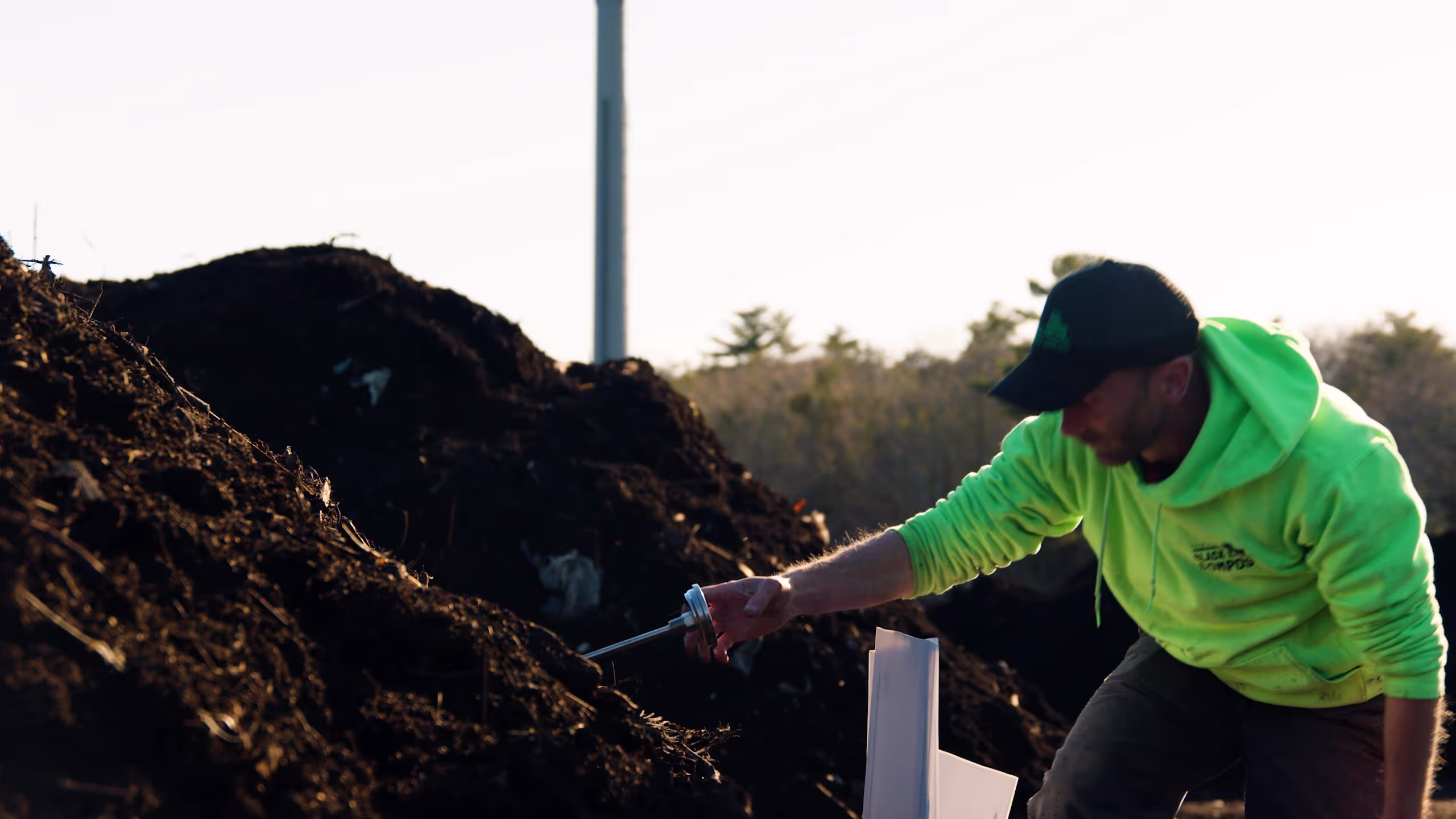 Person in neon green hoodie and black cap inserting a probe into a dark compost pile outdoors.