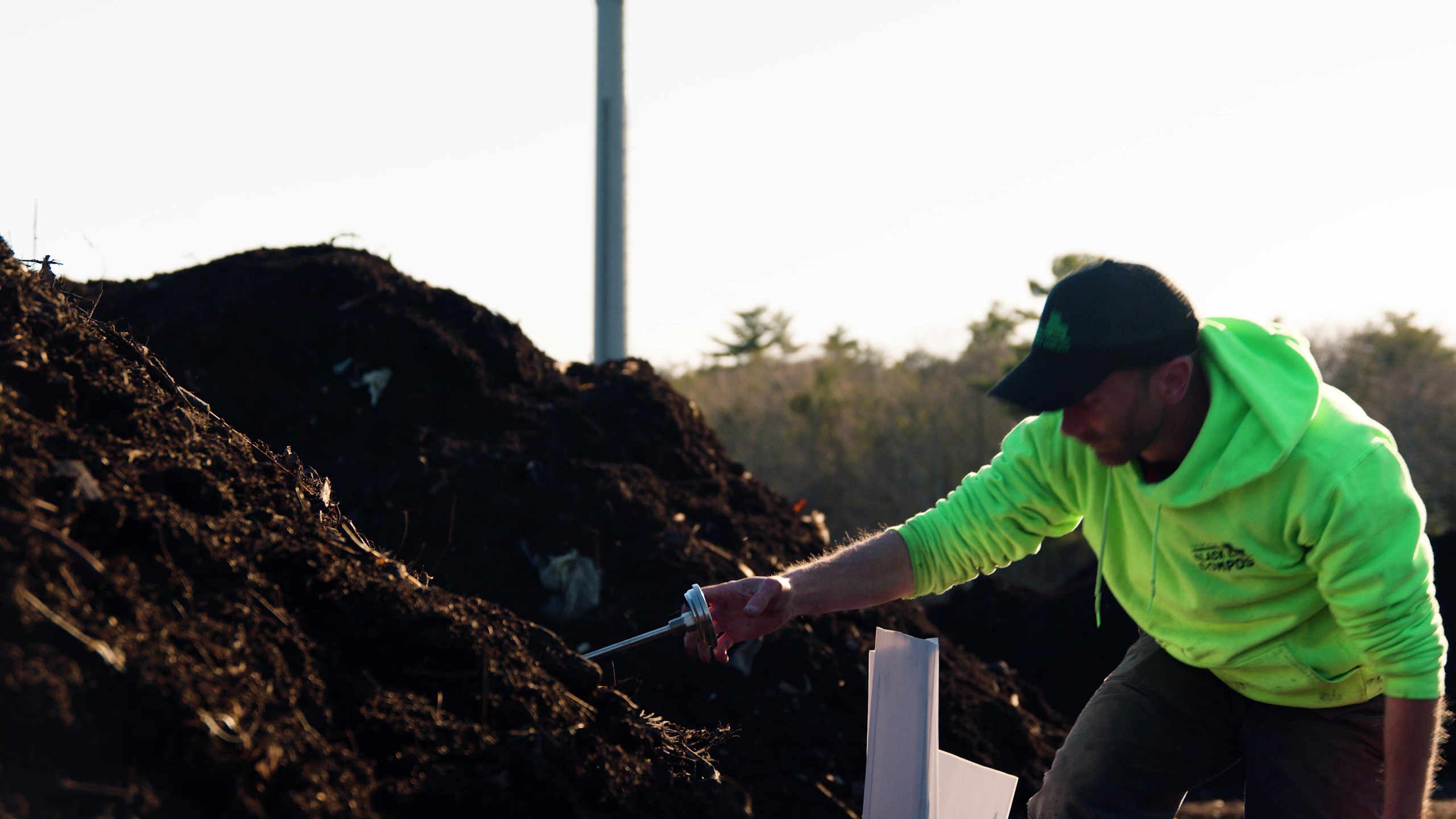 Person in neon green hoodie and black cap inserting a probe into a dark compost pile outdoors.