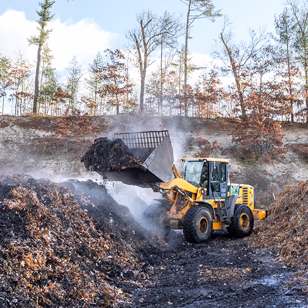 Yellow front loader moving steaming compost or mulch in an outdoor area with trees in the background.