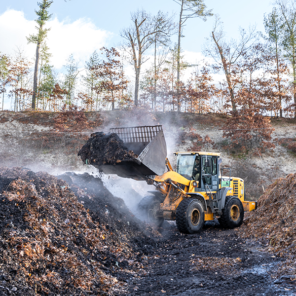 Yellow front loader moving steaming compost or mulch in an outdoor area with trees in the background.