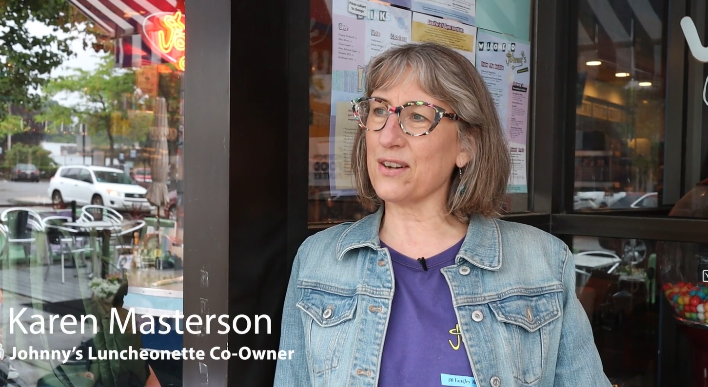 Woman with glasses and gray hair wearing a denim jacket and purple shirt stands outside a cafe with menu posted on the window behind her.