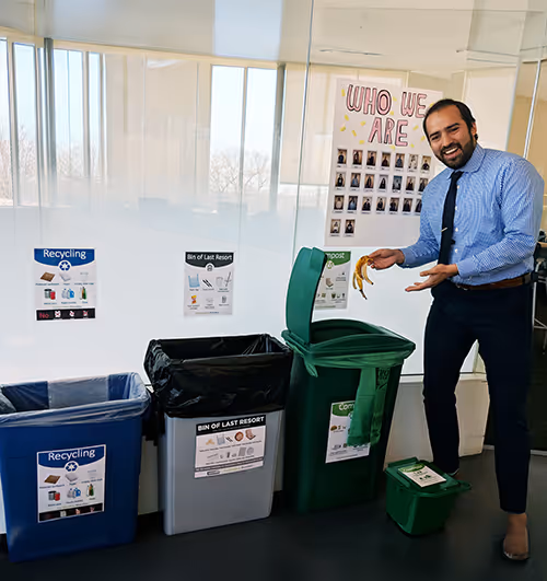 Man in office throwing a banana peel into a green compost bin next to recycling and trash bins.