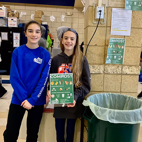 Two smiling girls standing indoors next to a green compost bin, holding a sign listing compostable food scraps and items.