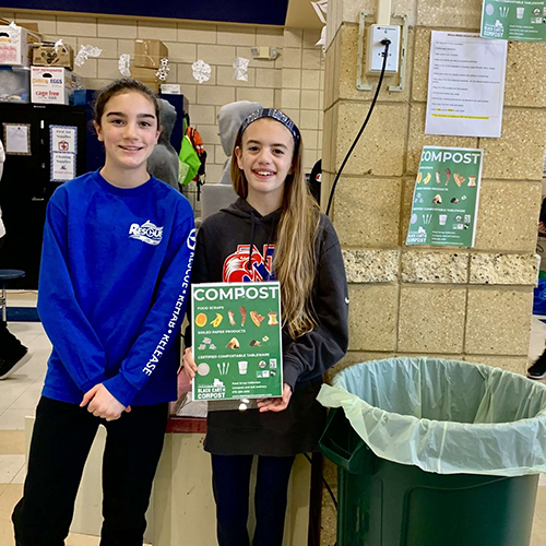 Two smiling girls standing indoors next to a green compost bin, holding a sign listing compostable food scraps and items.