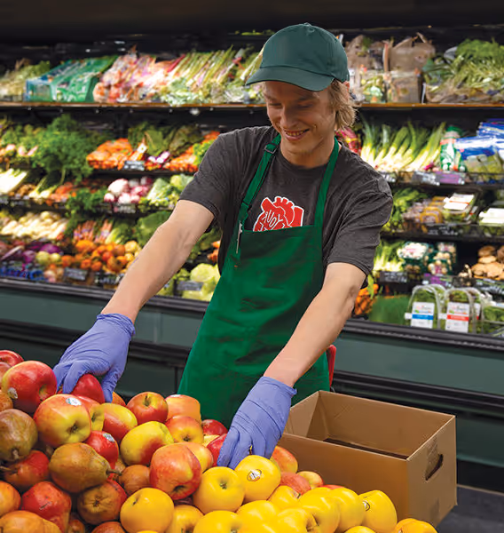 Grocery store worker in green apron and cap sorting apples into a box in the produce section.