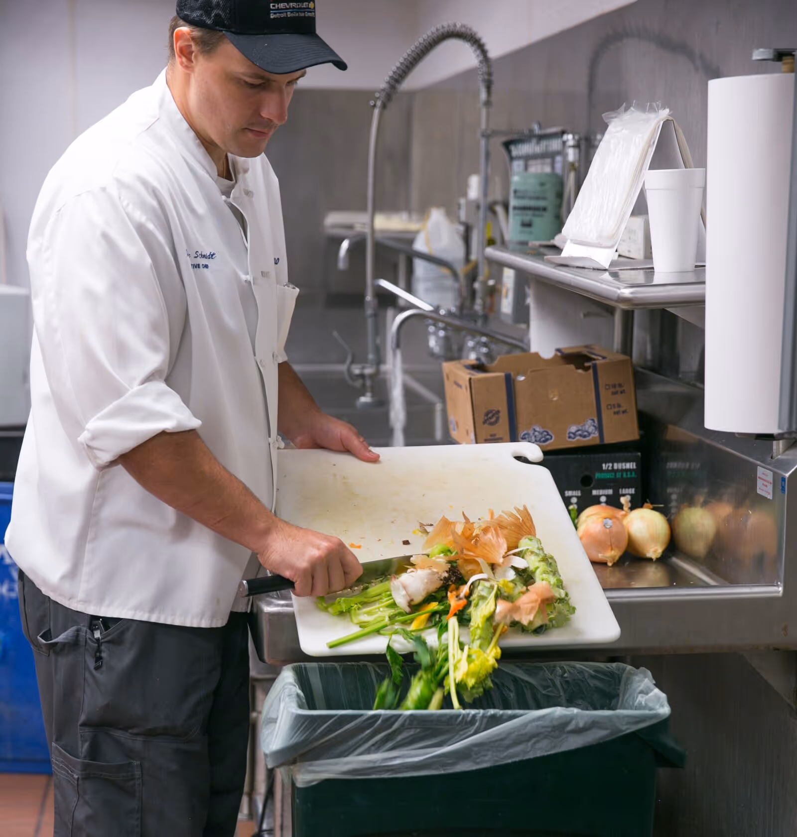 Chef in white coat scraping vegetable scraps from a cutting board into a compost bin in a commercial kitchen.