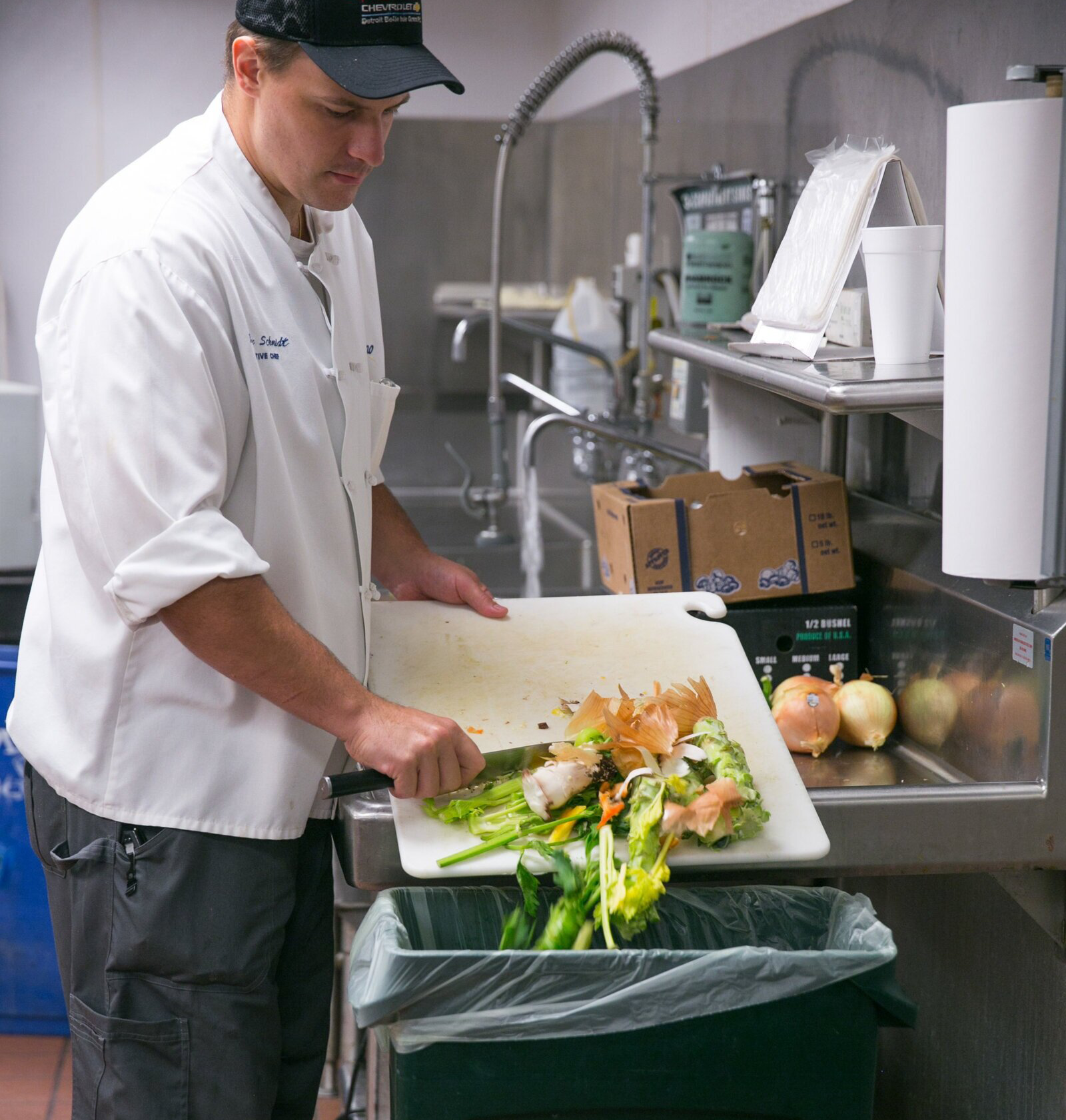 Chef in white coat scraping vegetable scraps from a cutting board into a compost bin in a commercial kitchen.