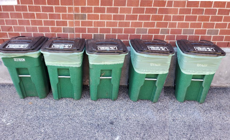 Five green recycling bins with black lids lined up against a red brick wall on a concrete surface.