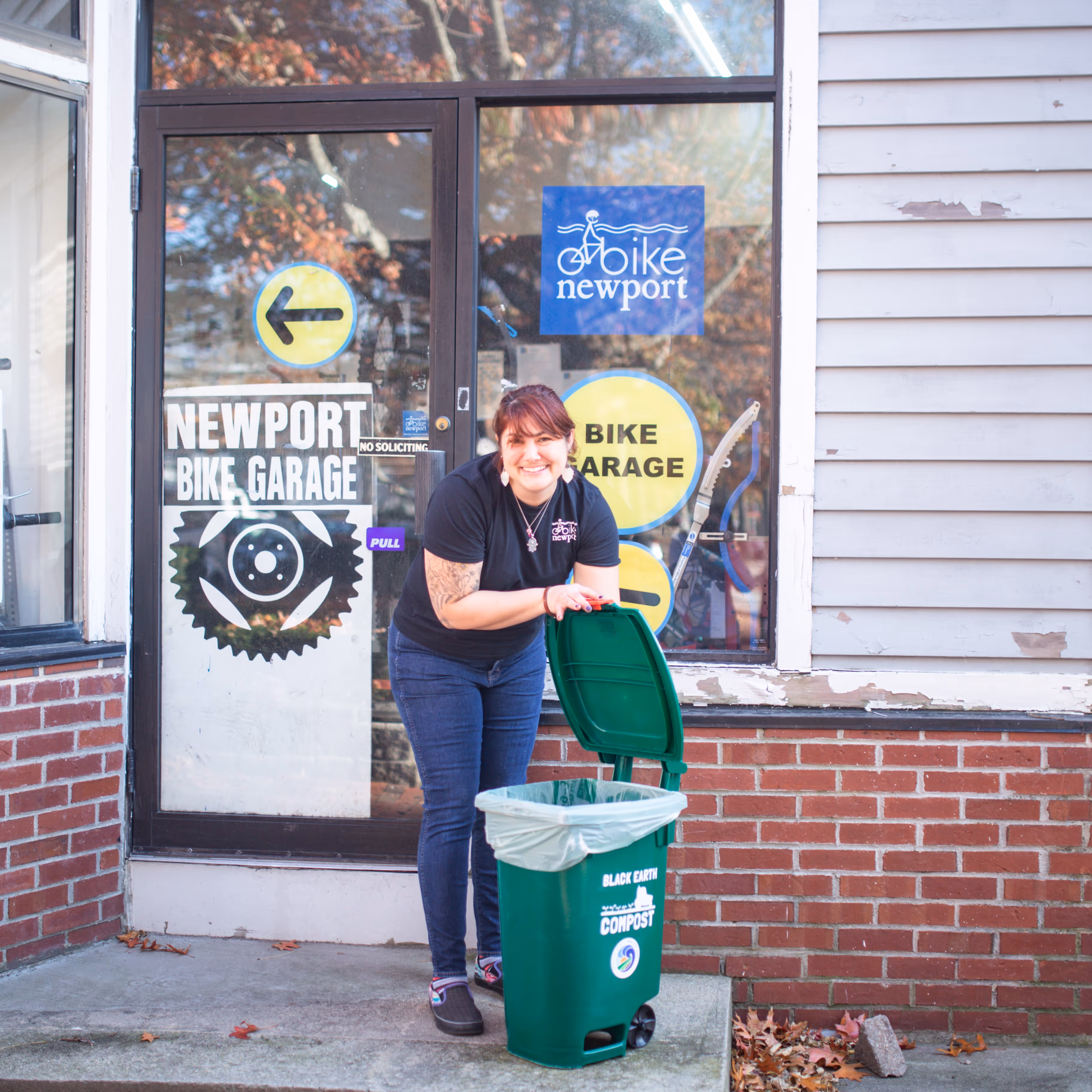 A woman smiling and holding open the lid of a green compost bin outside the Newport Bike Garage entrance with brick and siding walls.