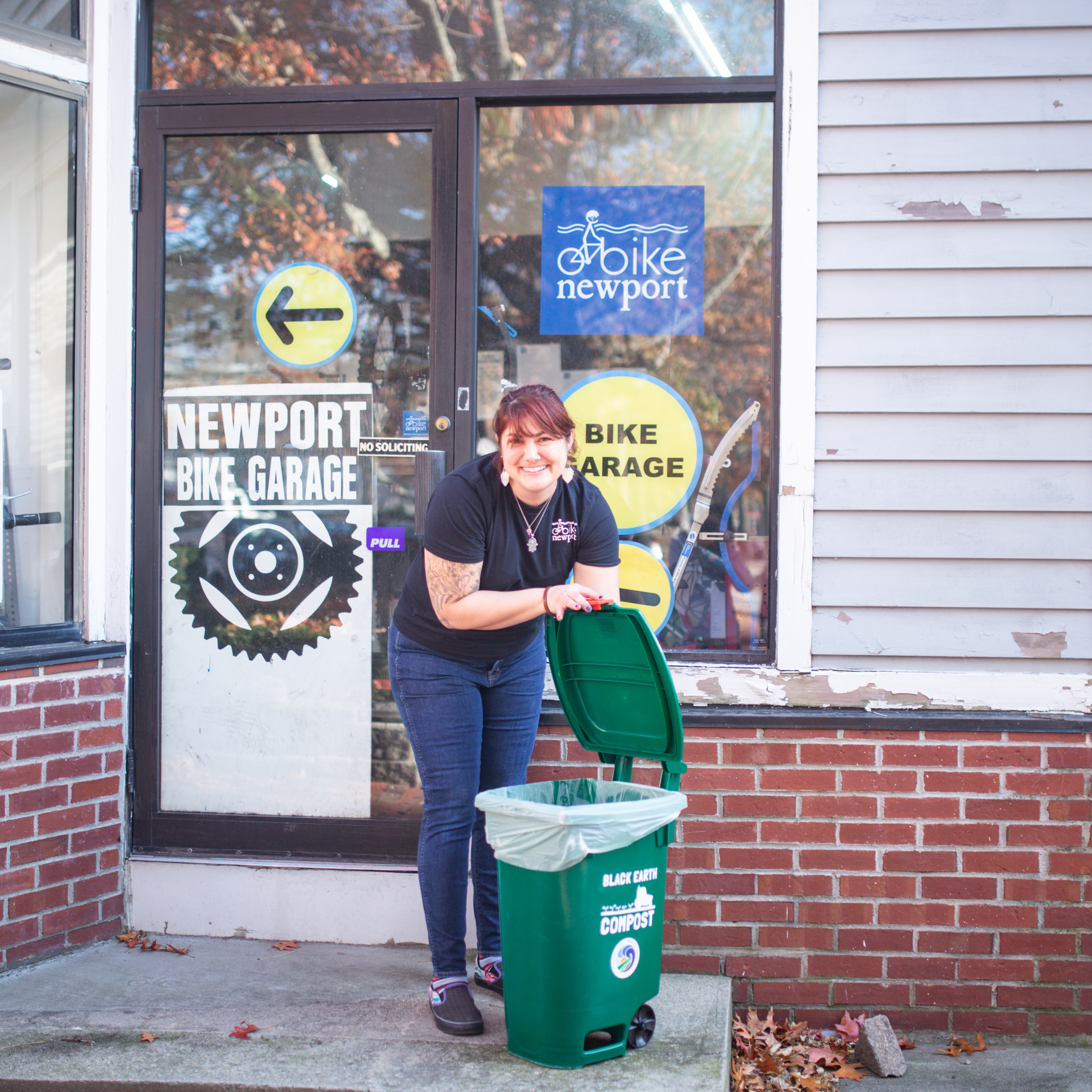 A woman smiling and holding open the lid of a green compost bin outside the Newport Bike Garage entrance with brick and siding walls.