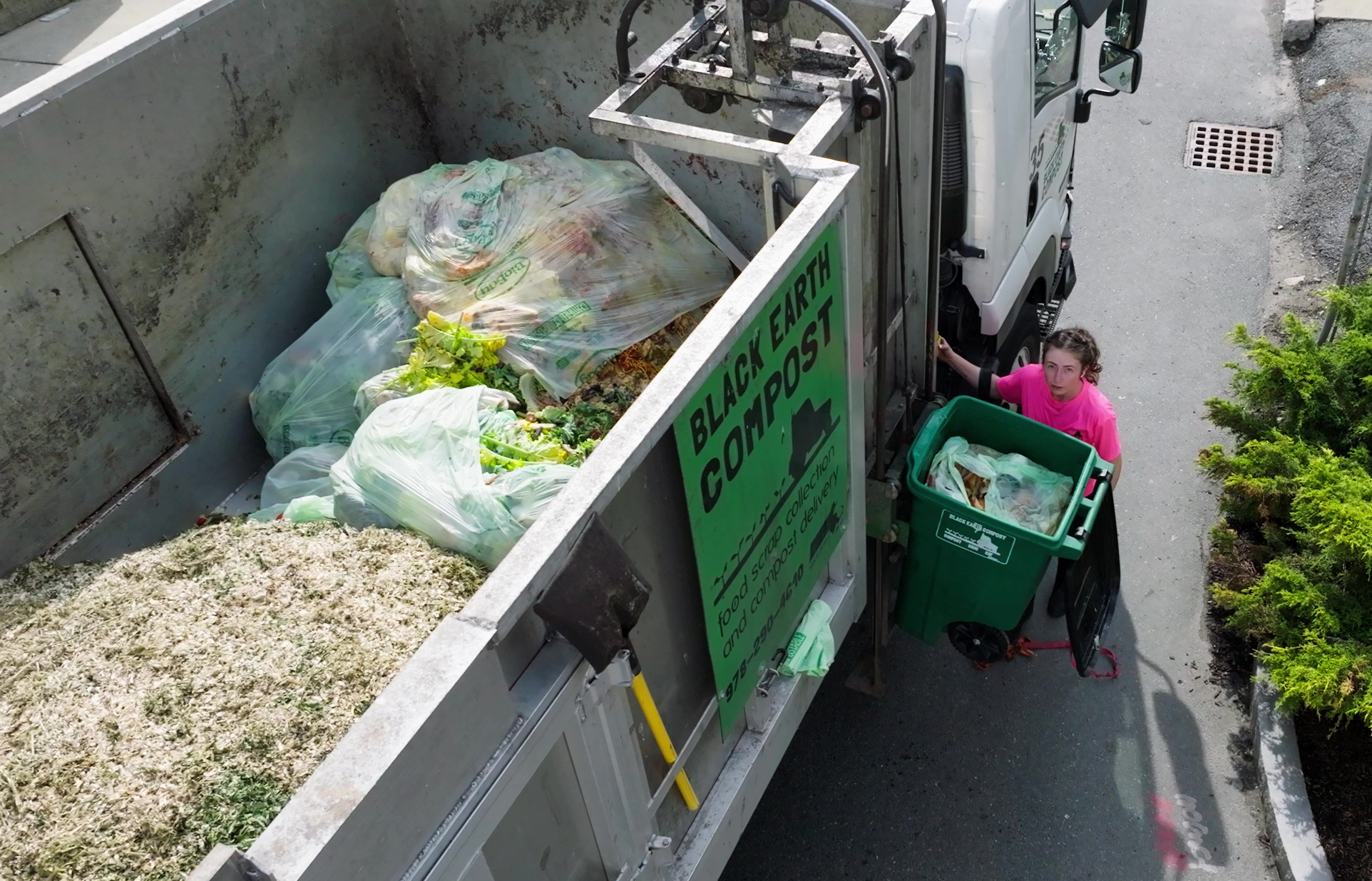 A woman in a pink shirt empties a green compost bin into a Black Earth Compost truck filled with bags of food scraps and compost.