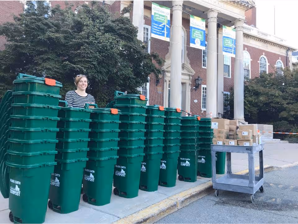 A woman stands behind a row of green compost bins labeled 'Black Earth Compost' stacked in front of a large brick building with pillars.