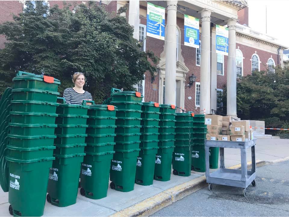 A woman stands behind a row of green compost bins labeled 'Black Earth Compost' stacked in front of a large brick building with pillars.