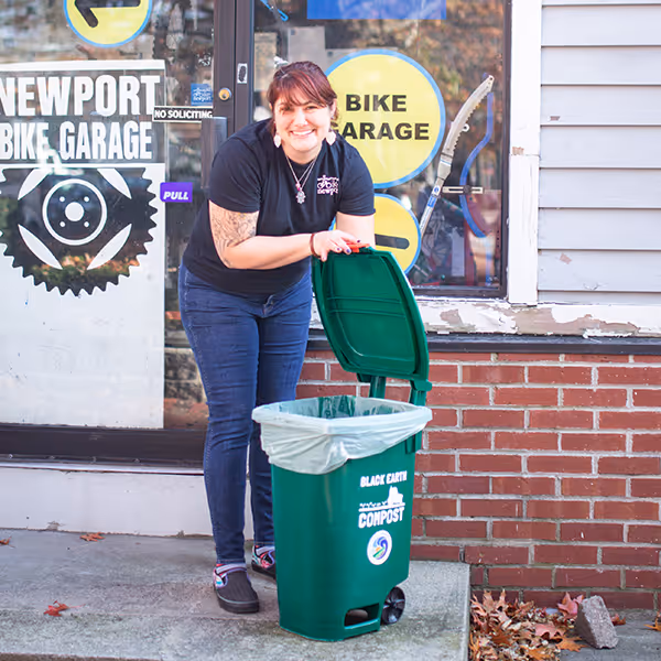 Woman standing outside Newport Bike Garage holding open the lid of a green compost bin.